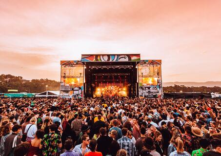 Music festival with a large crowd facing the stage. The stage is lit up with golden light. The sky behind the stage is turning red for the sunset. 