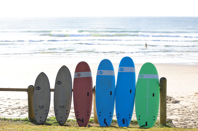 Surfboards and paddle boards lined up against a wooden fence, with the beach and ocean visible in the background.