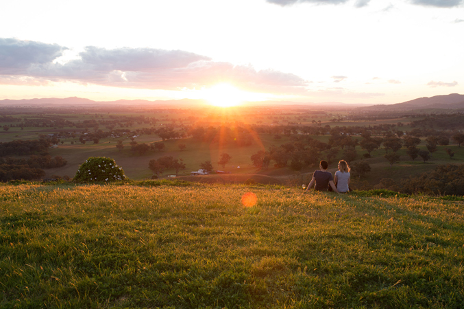 A couple sitting in a field of yellow flowers, watching the sunset over rolling hills in the distance