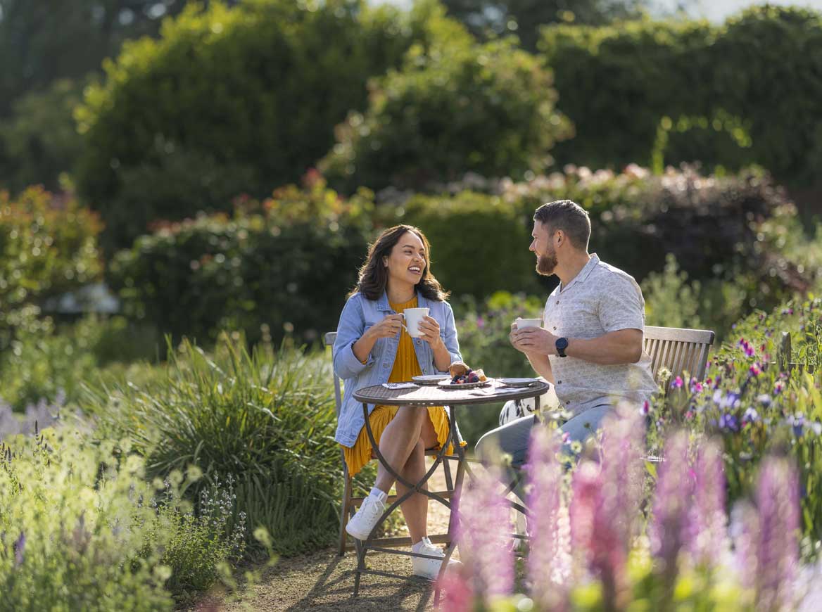 A couple sitting at an outdoor garden table, holding tea cups and laughing together.