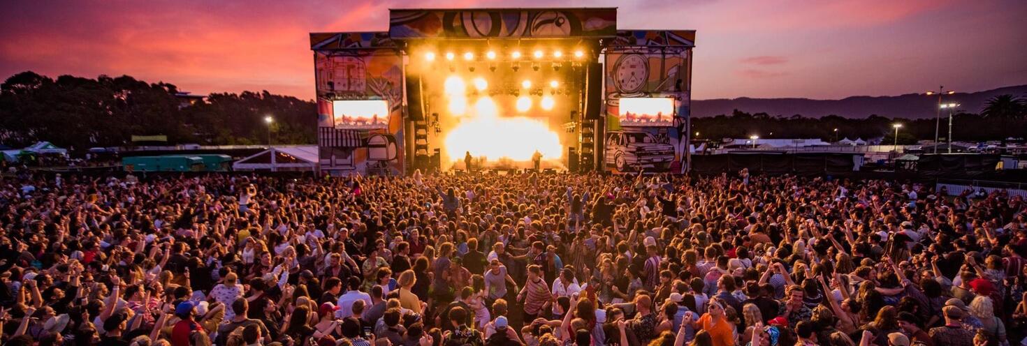 Large crowd of people standing in front of an outdoor concert stage. The sky is purple and pink tones at sunset. The stage has bright yellow lights beaming over the crowd.
