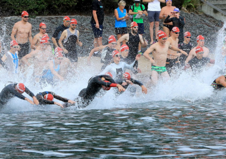 Many people are diving or running into the ocean with red swimming caps on. Most participants are wearing speedos or wetsuits. 